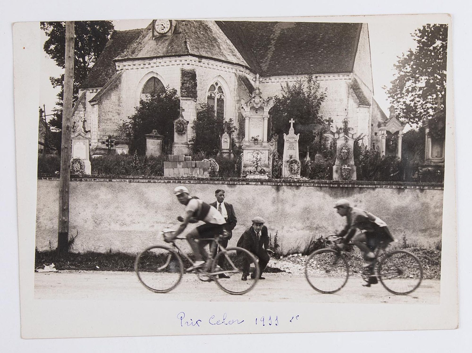 1933. Deux coureurs cyclistes participant au Prix Celor passent devant une église. Lieu non identifié.
