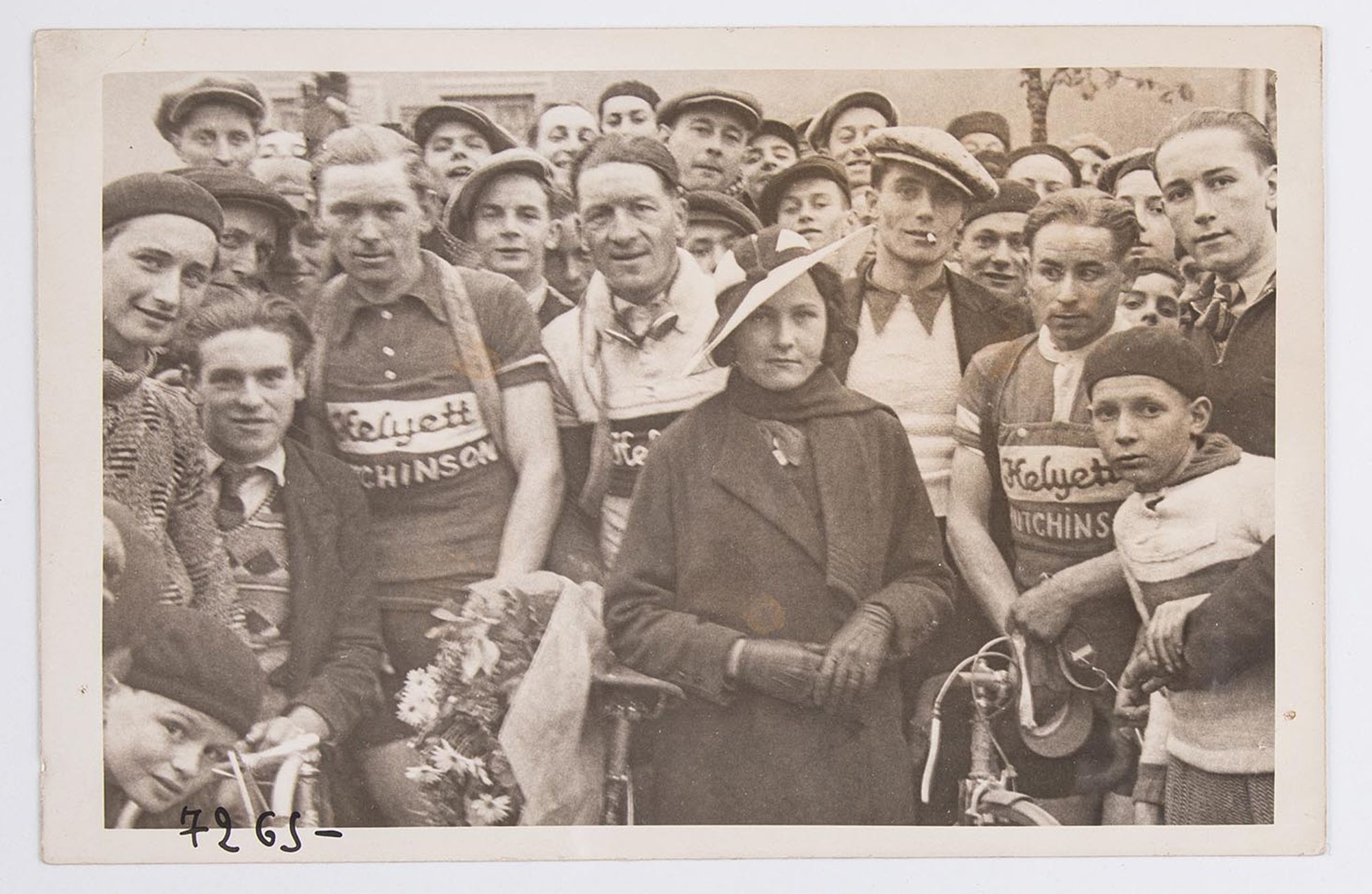 Photographie de groupe. Trois coureurs cyclistes posent au milieu d'un groupe de spectateurs. Ils entourent une jeune femme élégante. Lieu à identifier.