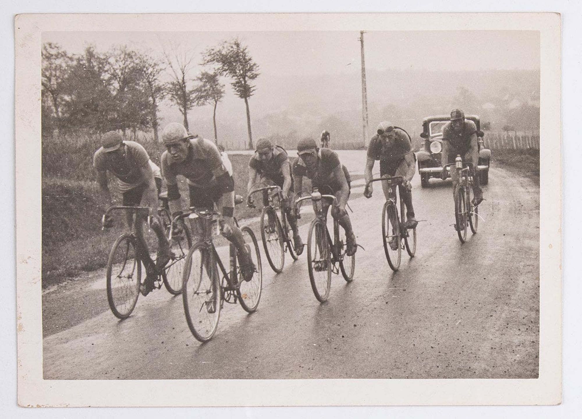 Groupe de coureurs cyclistes à l'assaut d'une côte, sous la pluie. Lieu à identifier.