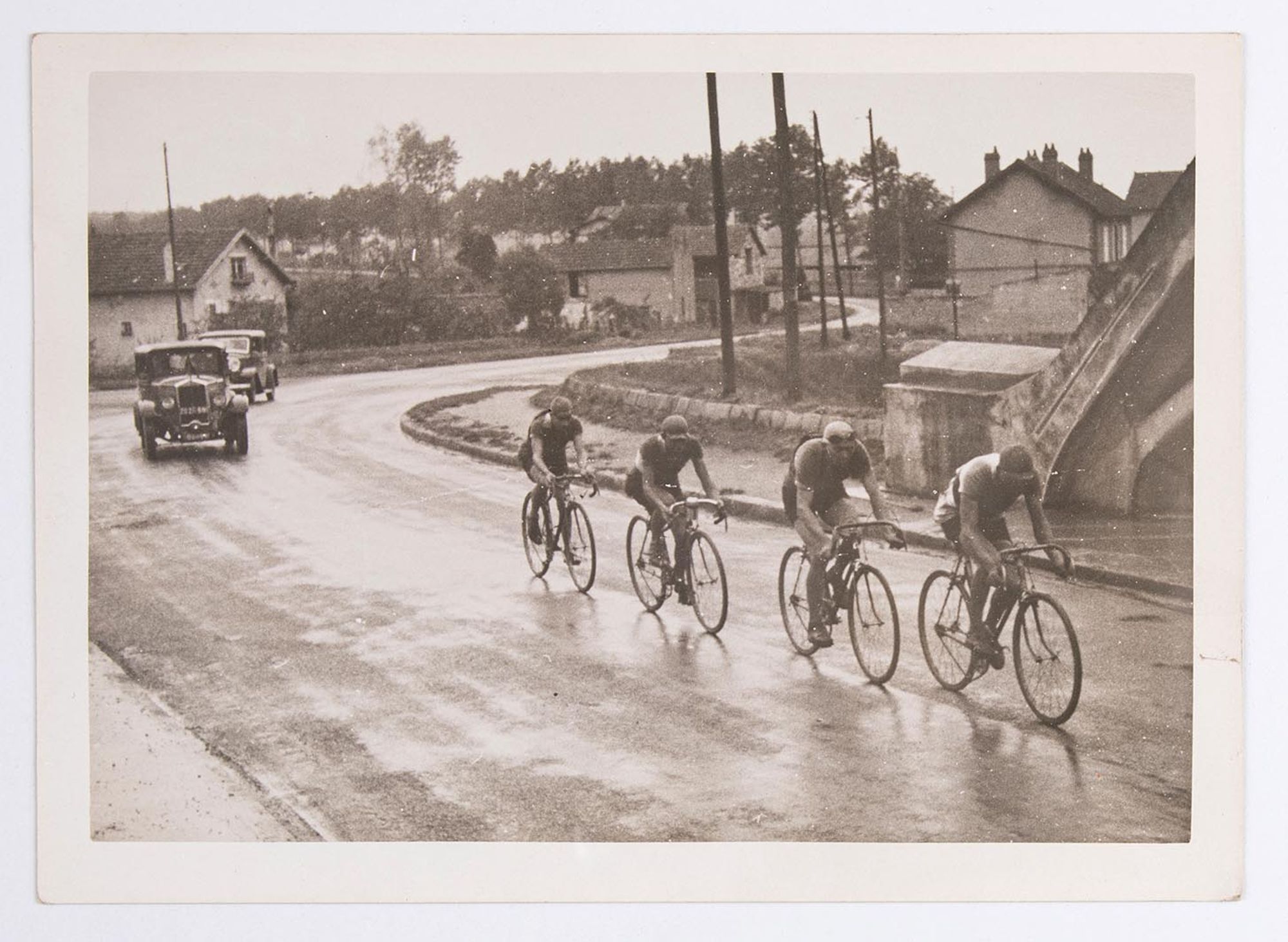 Quatre coureurs cyclistes affrontent une côte, au milieu des maisons. Course et lieu à identifier.