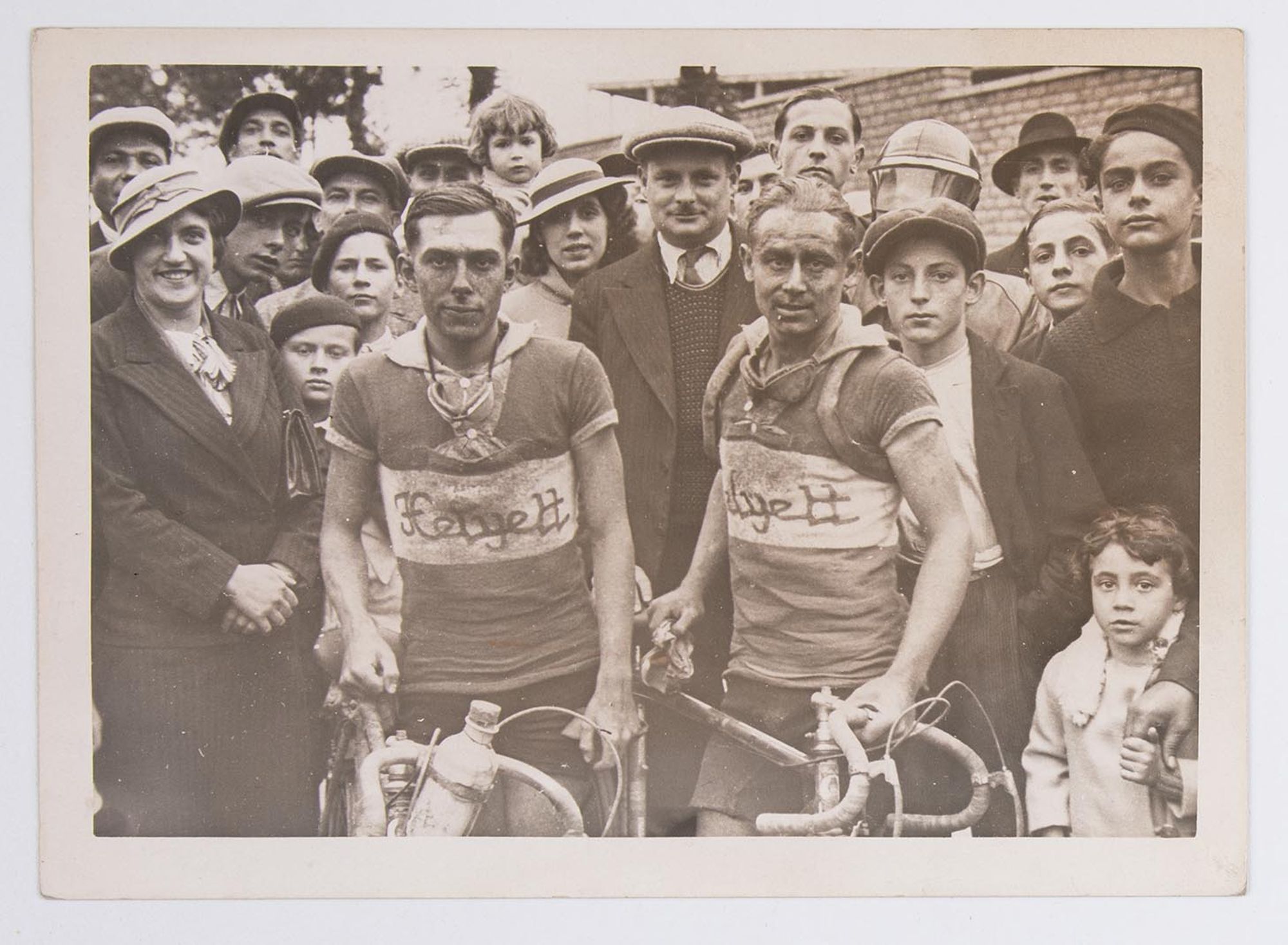 Portrait de groupe : deux coureurs cyclistes, portant un maillot Helyett, posent au milieu des spectateurs, à la fin d'une course. Course et lieu à identifier.